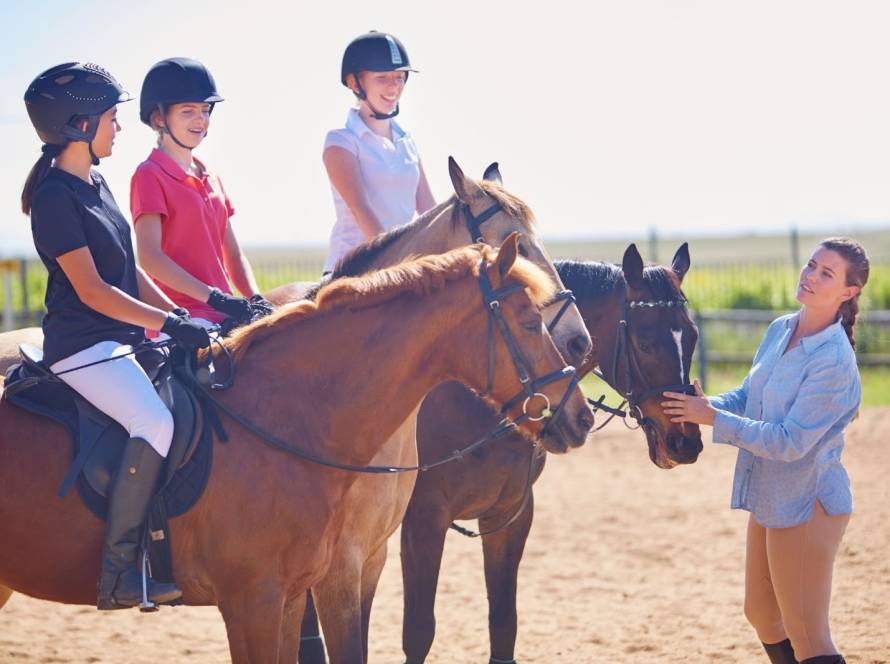 A female coach giving instructions to the girls during a horseback riding lesson in an outdoor riding ring